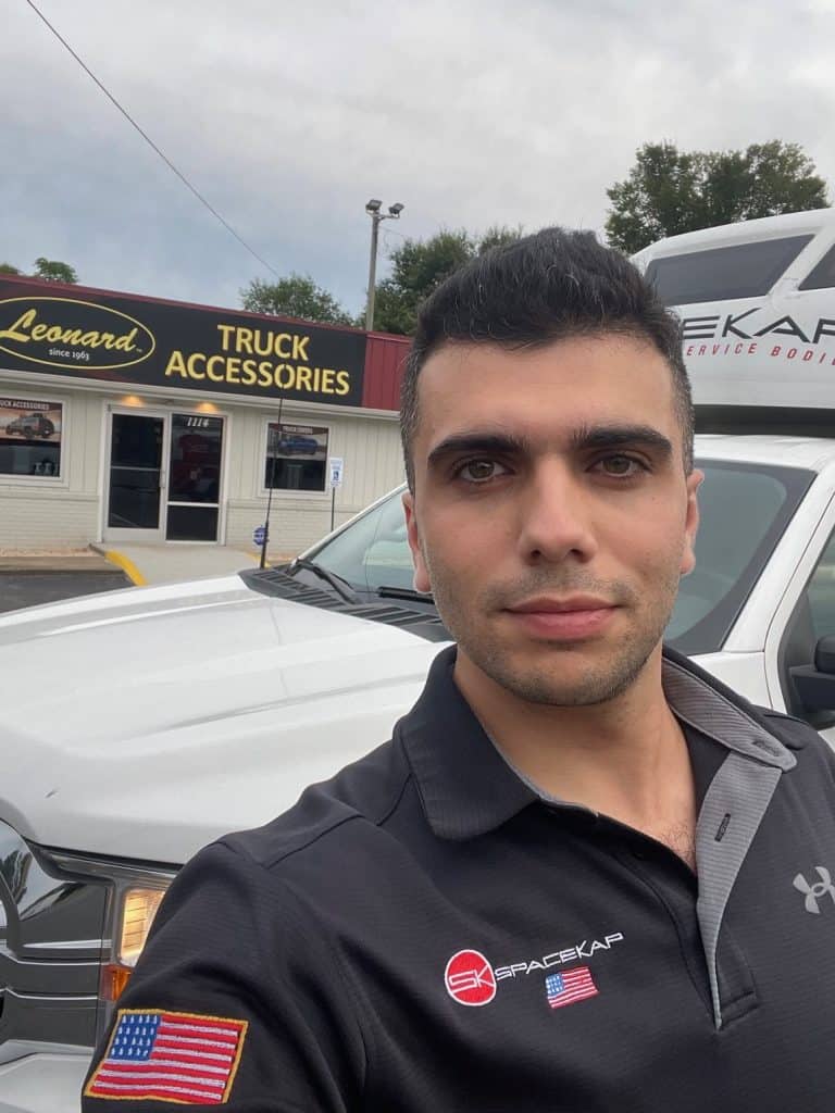 Homme prenant un selfie devant un camion blanc, avec un bâtiment de magasin d'accessoires pour camion en arrière-plan