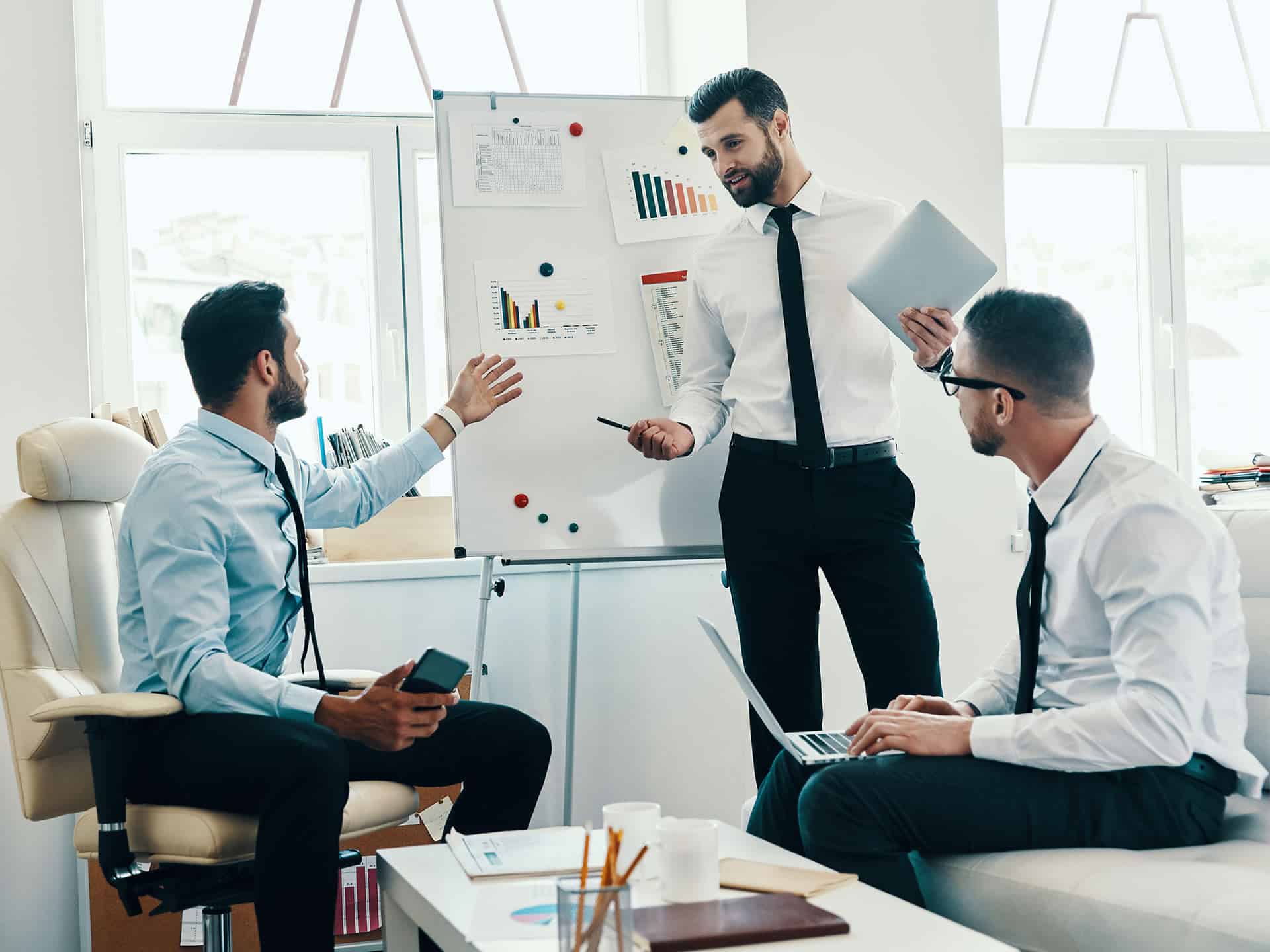Trois hommes en chemise et cravate discutant devant un tableau blanc avec graphiques